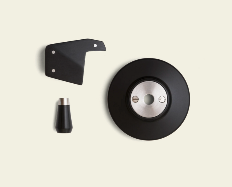 Set of mechanical components including a black disc, a smaller black piece, and a cylindrical component on a beige background.